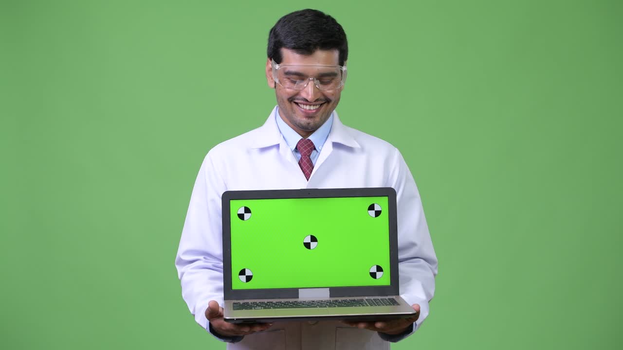 Young handsome Persian man doctor wearing protective glasses showing laptop
