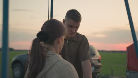 man prepares for flight by removing gas cylinder wrapped in red cloth from hot air balloon basket while woman in background opens car trunk during sunset on grassy field near parked vehicle