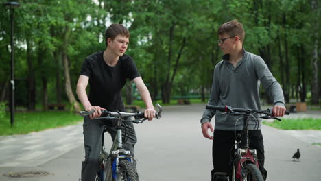 two friends on bicycles interact in a park setting, one in a grey top is waiting while his friend rides up to greet him with a knuckle bump, the background is blurred, showcasing lush green trees