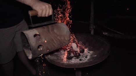 man empties hot charcoal on campsite grill with red embers rising up