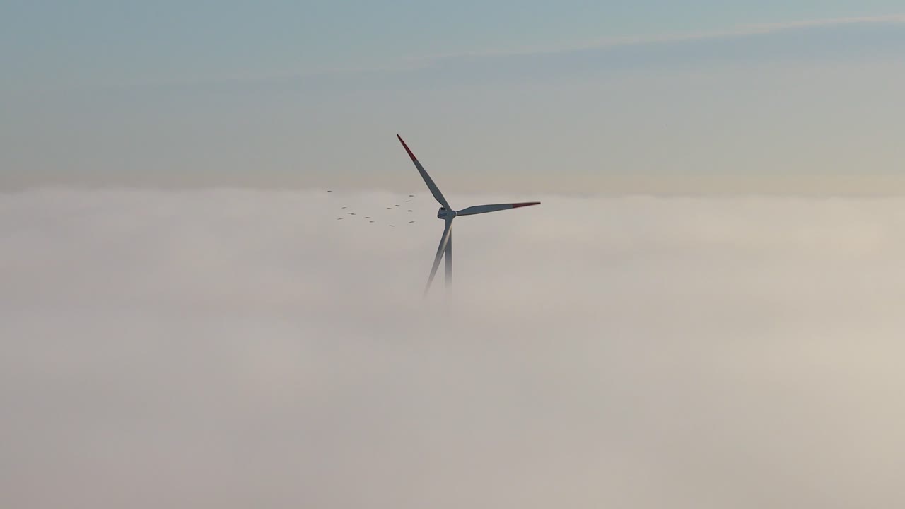 Veduta aerea cinematografica di una turbina eolica che fa girare le sue pale sopra un denso mare di nebbia durante una splendida alba a Danzica, Polonia.