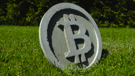 close up or closeup pullout footage of a concrete gray colored bitcoin cryptocurrency sculpture coin or a medallion during a sunny summer day in a garden with mowed lawn and a hedge in the background
