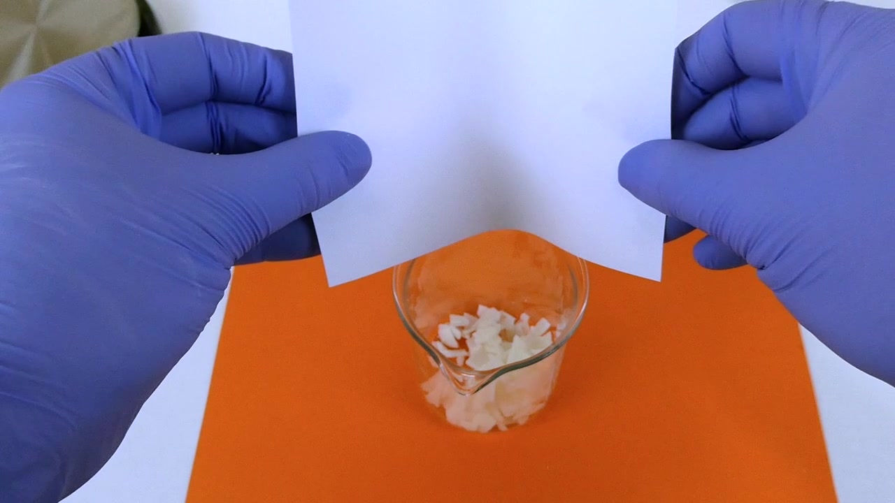 Científico en un laboratorio vertiendo escamas blancas de hidróxido de sodio de un papel en un vaso de precipitados de vidrio