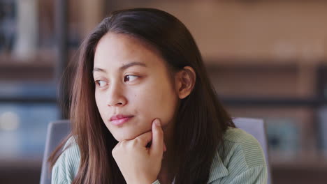 millennial asian female creative looking way in contemplation, resting chin on hand, close up