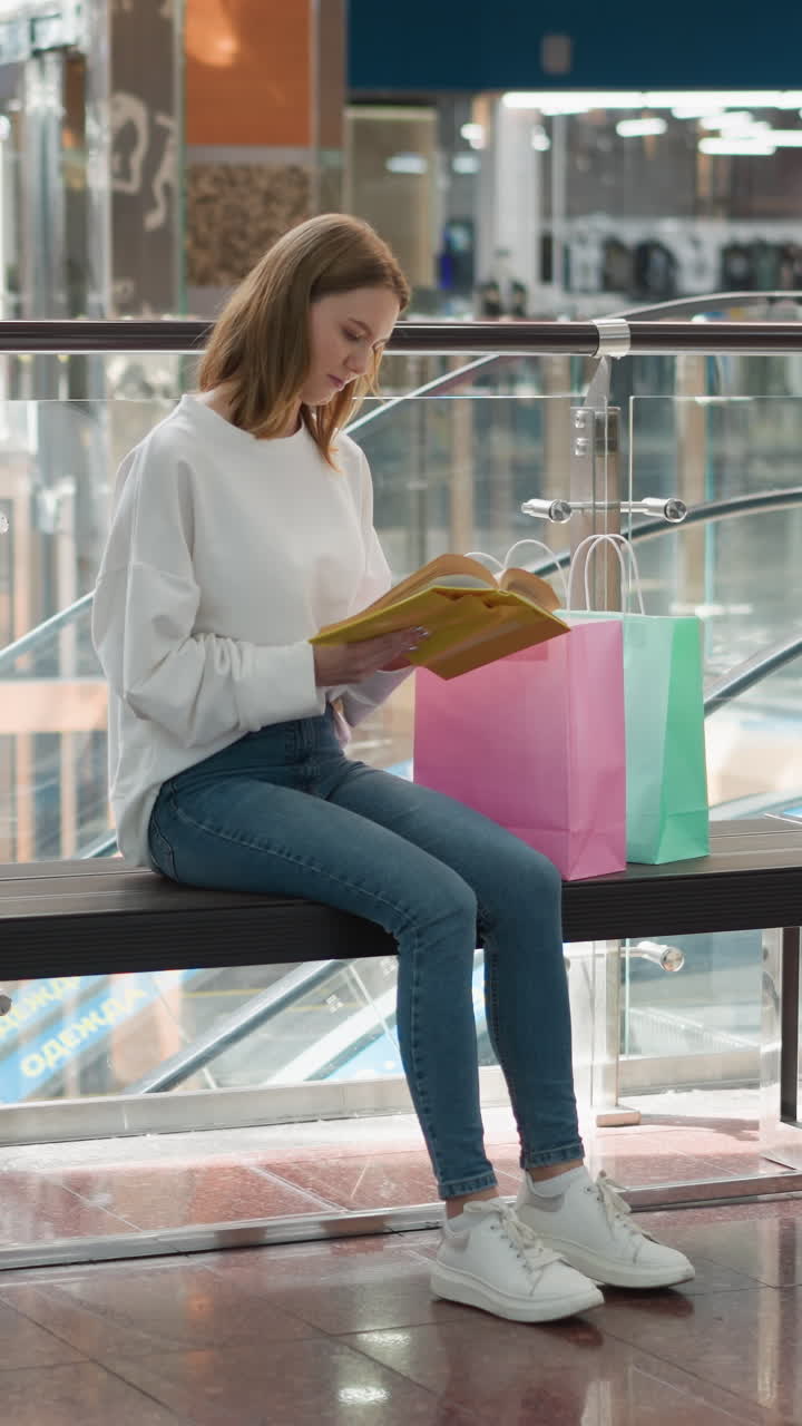 mujer joven sentada en un banco en un vibrante centro comercial leyendo un libro amarillo mientras balancea la pierna lentamente, bolsas de compras de colores visibles, barandillas de vidrio modernas y ascensor en movimiento