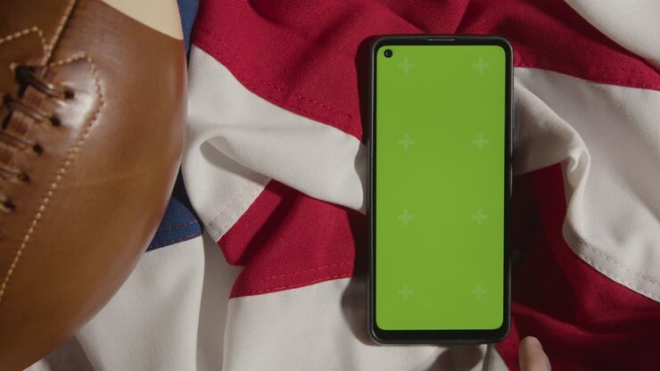 Ripresa in studio dall'alto di una palla da football americano su una bandiera a stelle e strisce con una persona che utilizza un telefono cellulare con schermo verde.