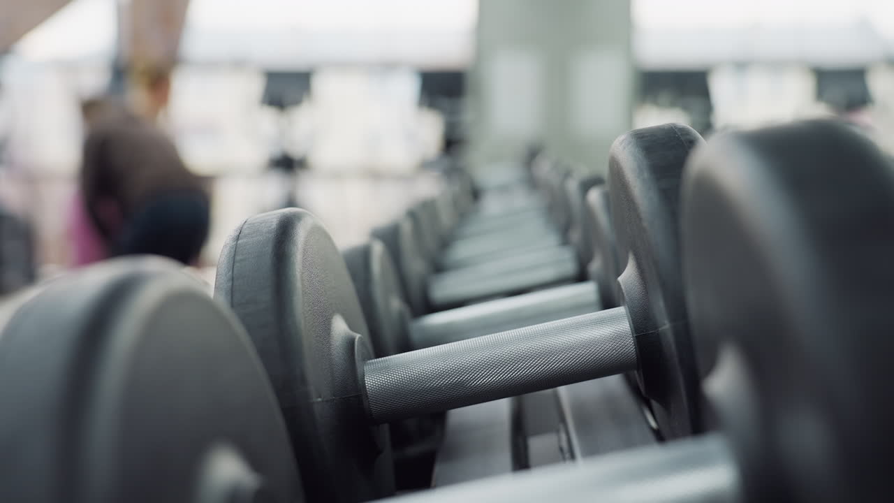 Dumbbells neatly lined up on rack in bright gym with blurred exercisers in background, showcasing sleek equipment, modern fitness space, focus on weights and workout environment