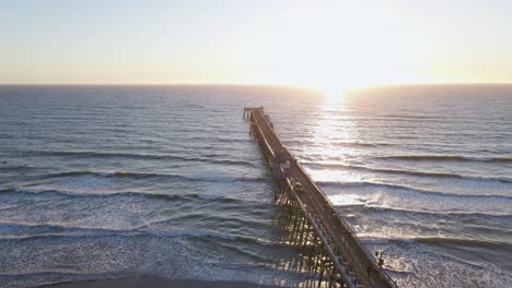 aerial view of pacific ocean with waves breaking into a long pier at sunset