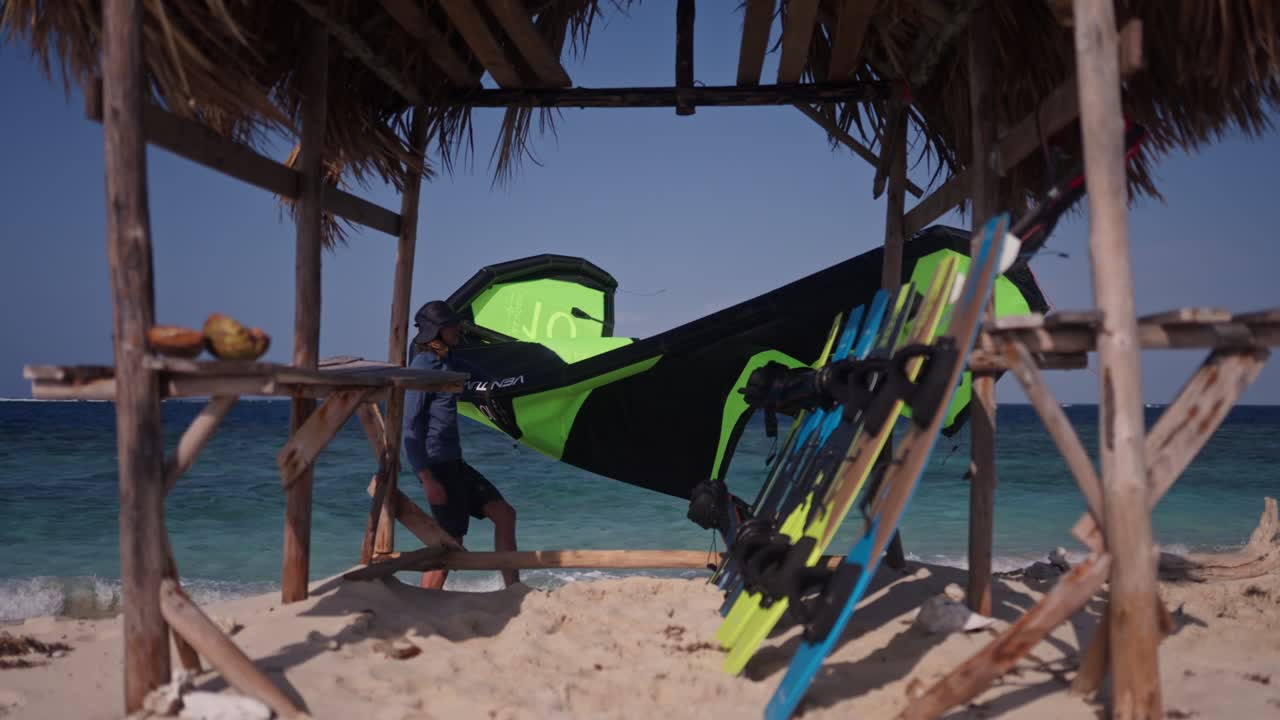 disparo medio estático de un kiteboarder sosteniendo una cometa caminando más allá de una pequeña cabaña abierta con tablas inclinadas hacia adentro y el océano en el fondo