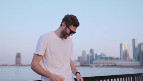 Happy-handsome-man-in-sunglasses-and-white-t-shirt-uses-a-watch-watch-looks-and-presses-his-finger-on-the-screen-standing-on-the-waterfront-in-summer-against-the-city-and-buildings