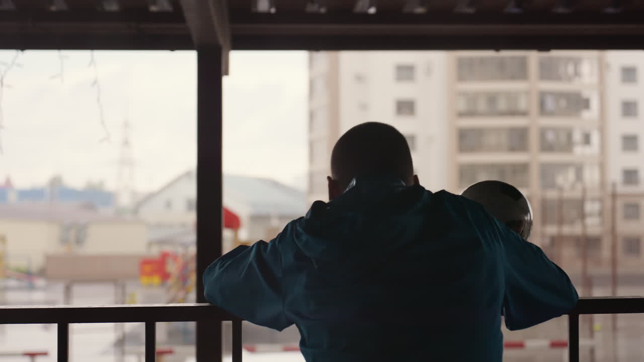 man on balcony watching rain, cradling football and staring at blurred city skyline through droplets, contemplative solitude, muted colors, quiet longing and reflective pause in apartment terrace