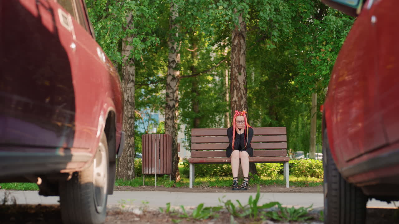 차들 사이에 있는 공원 벤치, 픽업을 기다리는 백인 통근자, 타이어가 보이는 낮은 도로 시점, 시계와 가방을 확인, 자작나무 덮개를 통과하는 여름 햇빛, 도시 대중교통 정차