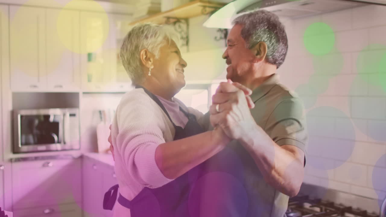 Animation of colourful spots over happy senior diverse couple dancing at home