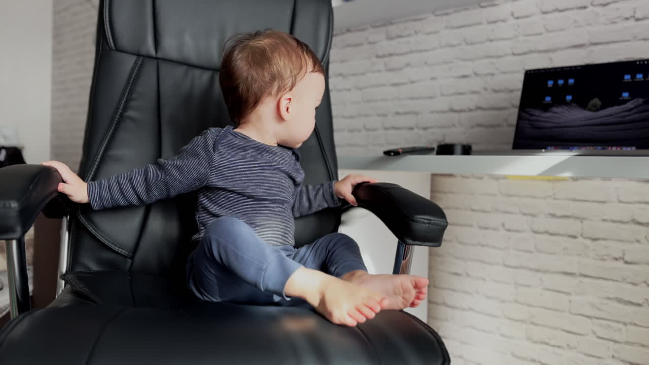 Little baby boss sitting comfortably in a big leather chair. Cute boy takes tv remote from a table.