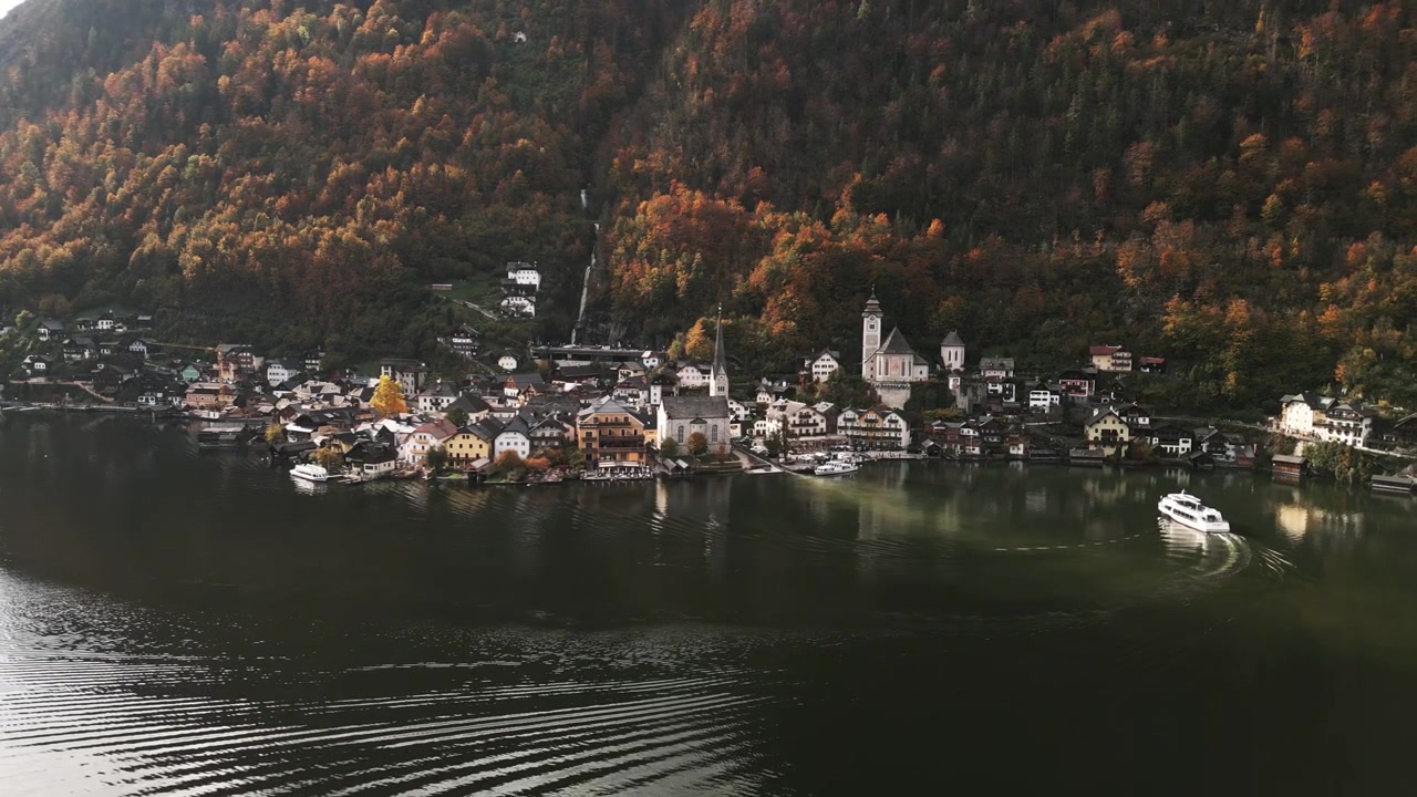 Hyggelig landsby beliggende ved en rolig sø, der viser farverige efterårstræer og charmerende bygninger. En båd glider hen over det grønne vand.