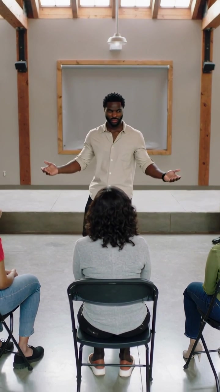 Vertical video: Descending off stage man addressing group on folding chairs in hall, rolled-sleeves