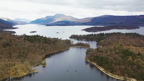 ángulo-Aéreo-De-Drones-De-Barcos-Que-Viajan-A-Través-De-Los-Estrechos-En-Loch-Lomond,-Escocia,-Con-Luss-Y-Ben-Lomond-En-El-Fondo-A-Principios-De-La-Primavera