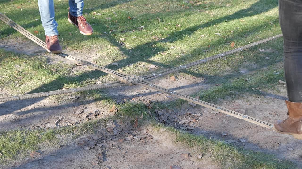 Man and woman playing on slackline balancing in front of each other. Two ropes cross each other in the center making a cross. Man balances on one foot then jumps down. Shadow projected on park grass
