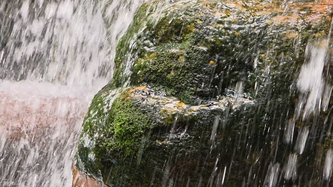 A powerful close-up shot of a waterfall, where clean water cascades and splashes over a large, ancient rock covered in green moss, creating the illusion of a natural face profile.