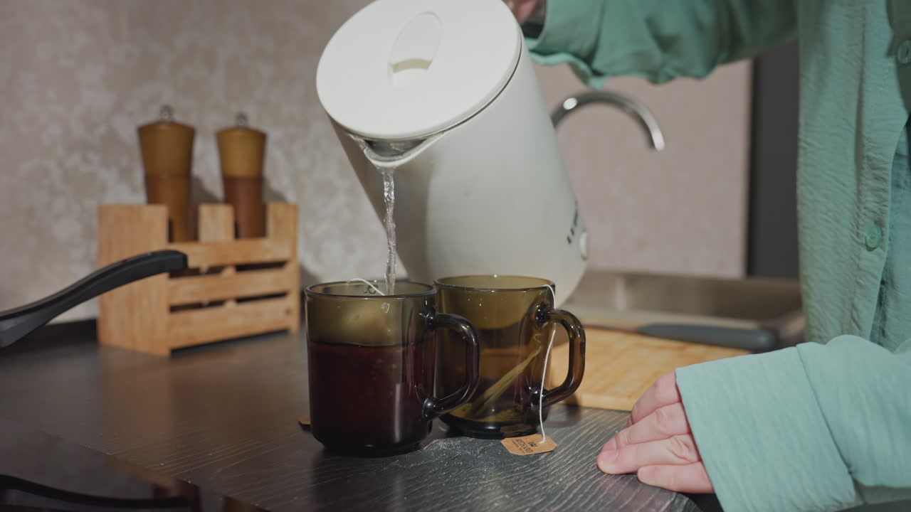 Close up of person in green pajamas pouring hot water from electric kettle into two transparent glass cups with tea bags on kitchen counter, wooden cutting board and spice jars visible in background