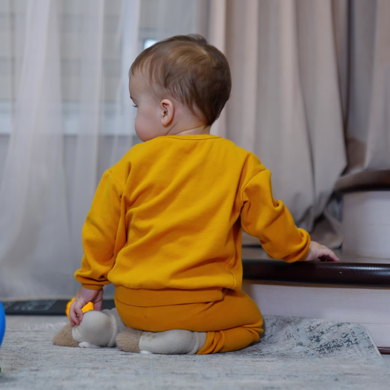 Lovely toddler in orange sport suit sits on the floor near the stairs. Kid throws the toy and watches it roll