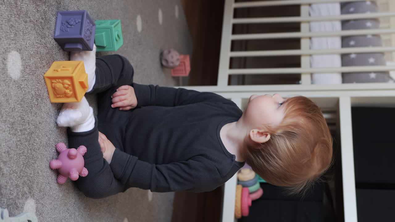 Vertical video of curious baby girl sitting on bedroom floor, playing with toys