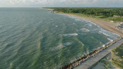 widok z lotu ptaka na betonowe molo w porcie liepaja, wybrzeże morza bałtyckiego, słoneczny letni wieczór, światło złotej godziny, pluskające wielkie fale, szerokie ujęcie drona poruszające się do przodu