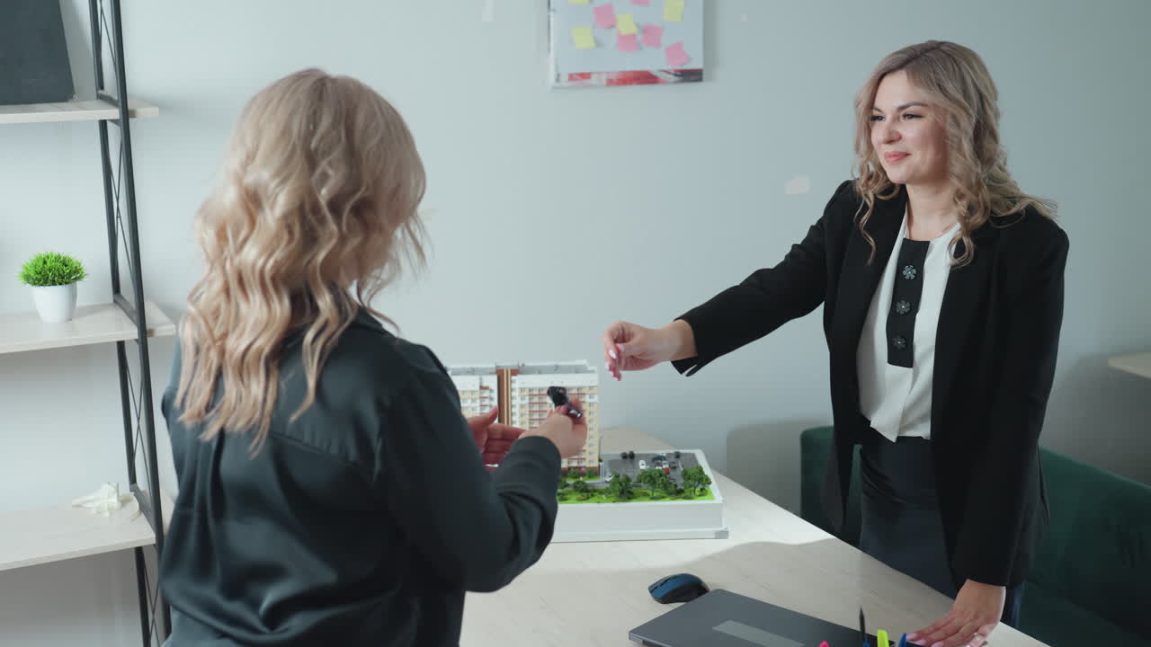 saleslady rises to present key near detailed building miniature as client stretches forth hand to receive key and they exchange handshake with warm demeanor beside pen holder on office table