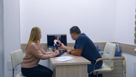 medic points at the skeleton image on the screen. doctor explains the health problem to a blonde female patient.