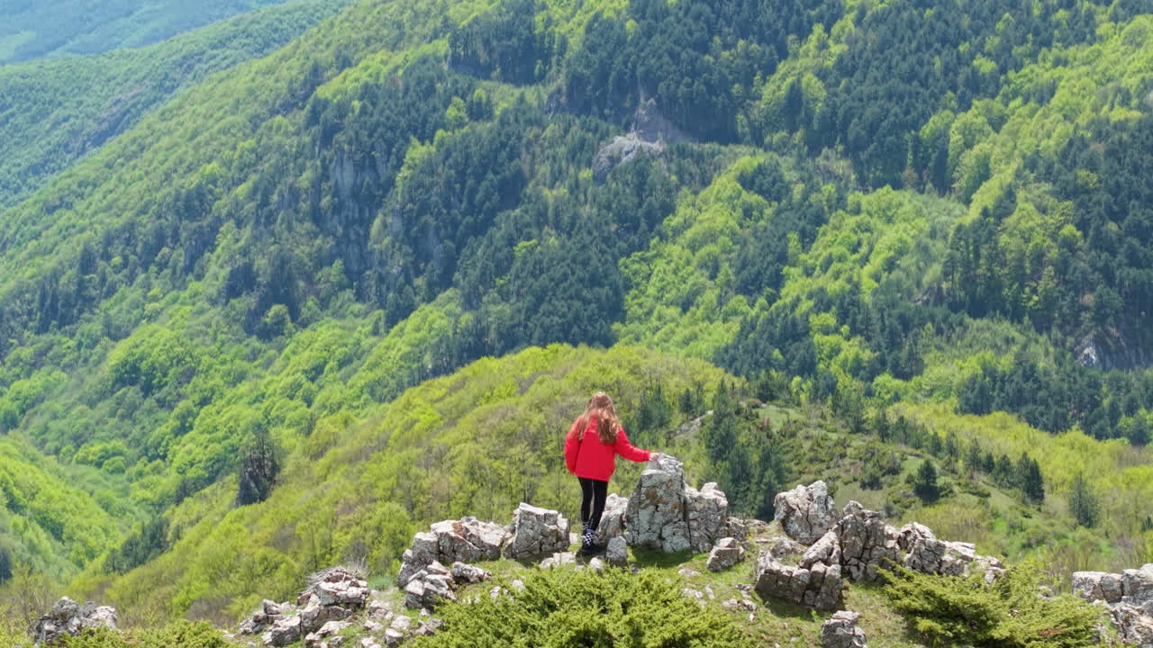 A solo female hiker enjoys a panoramic view over meadows, forests, and rolling hills at Beklemeto Pass, Bulgaria. The perfect scene of peace, freedom, and adventure in nature during springtime.