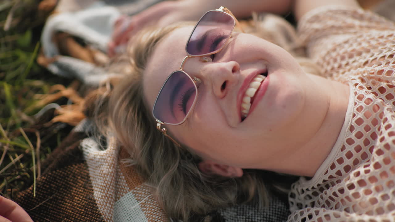 dama feliz con camiseta y gafas de sol acostada en una sábana al aire libre, sonriendo mientras toca la hierba, disfrutando del calor de la luz solar y el aire fresco en un entorno rural pacífico