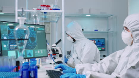 doctor in ppe suit working at pc while chemist using microscope