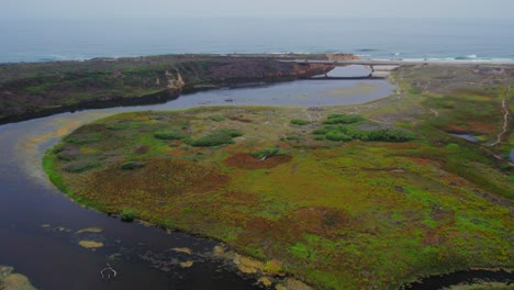 Rotating-drone-shot-of-a-winding-lagoon-connecting-to-the-sea-and-revealing-a-bridge-with-cars-crossing-in-the-background