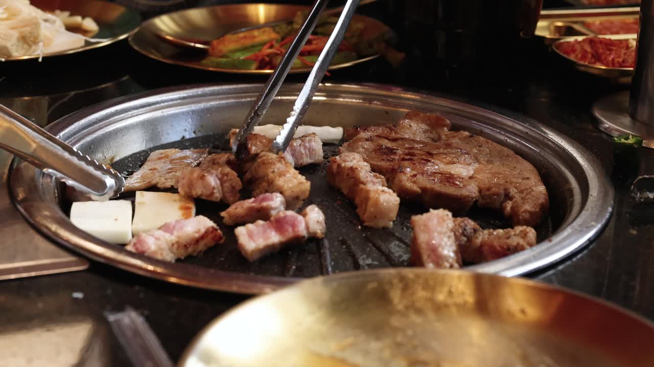 Tongs turn pork belly and meat slices on tabletop grill, warm lighting, restaurant setting, close-up