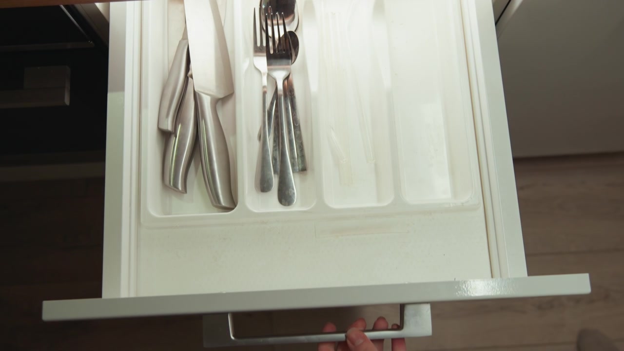 Hand in green sleeve pulls open wooden kitchen drawer to reveal white molded utensil tray holding neatly arranged forks, spoons, knives, with reflective metal surfaces and clean domestic storage