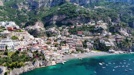 Amplia-Vista-Aérea-Que-Muestra-Positano-Rodeado-De-Aguas-Cristalinas-Con-Barcos,-Exuberante-Vegetación,-Casas-Costeras-Y-Un-Cielo-Azul-Claro-En-El-Fondo,-Costa-De-Amalfi,-Italia