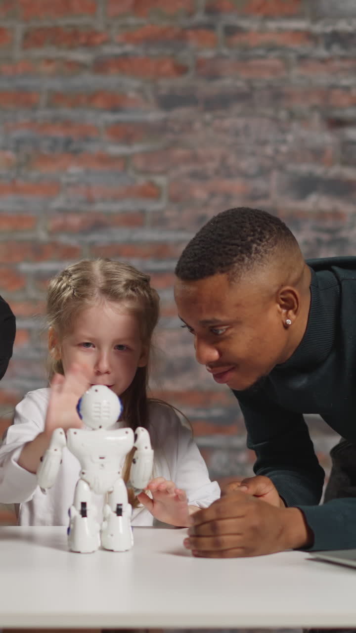 Little girl waves hand to control funny robot reaction learning engineering with positive Asian and African-American science teachers at table