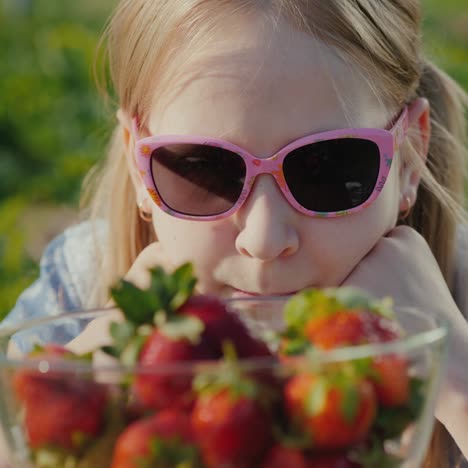 girl in sunglasses looks at a bowl of ripe strawberries