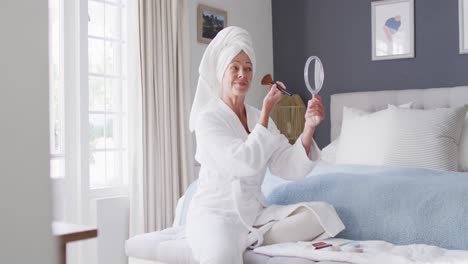 happy senior caucasian woman sitting on bed in bedroom, putting on make-up