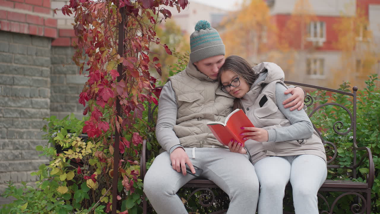 Bonding moment between lovers seated outdoor as man gently pats woman hand while she reads book in peaceful autumn setting surrounded by colorful foliage and gentle breeze swaying leaves