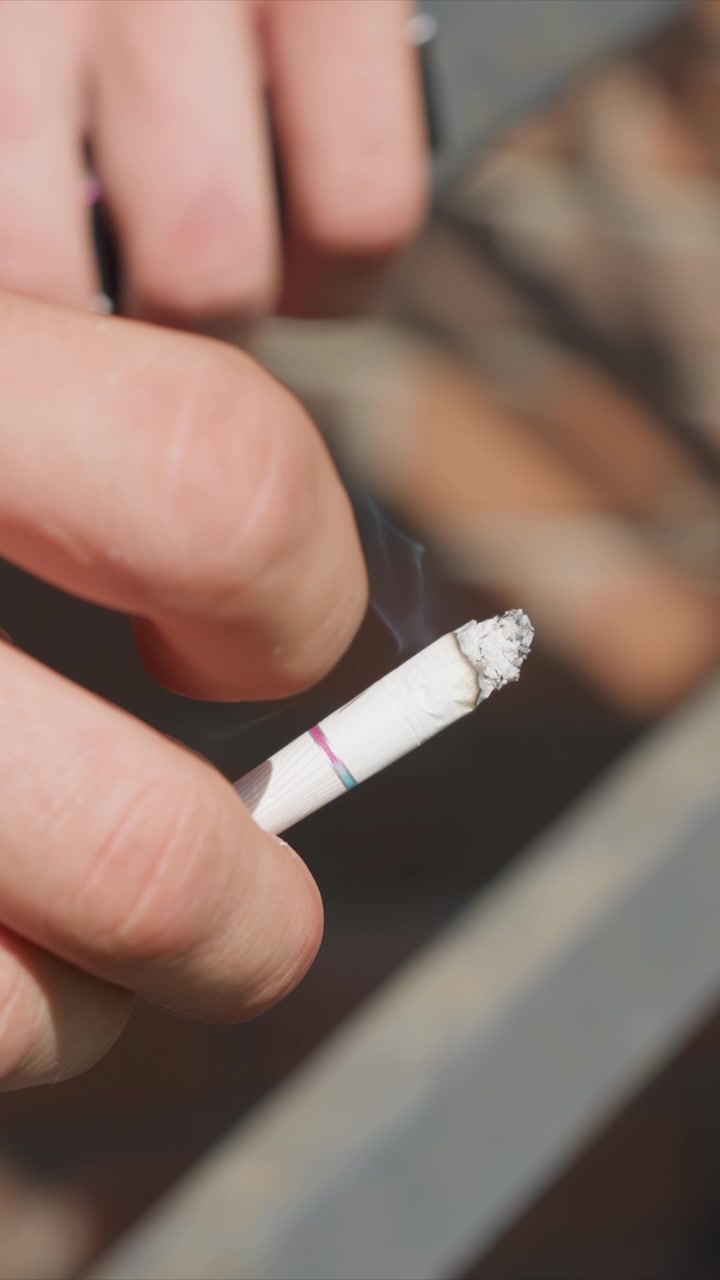 Primer plano de una mano sosteniendo y manipulando un cigarrillo sin encender con ceniza en la punta. El enfoque se mantiene en los dedos que sujetan el objeto cilíndrico blanco, mostrando un ligero movimiento mientras se gira o ajusta.