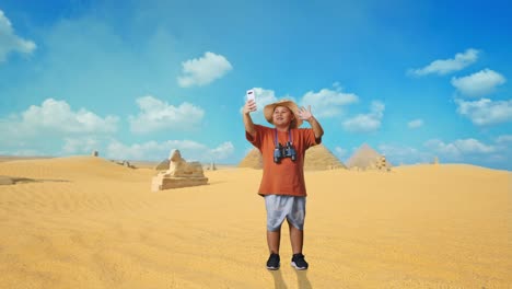 asian boy with a hat and binoculars waving hand having video call on smartphone while traveling in giza pyramid. boy researcher examines something, travel adventure, full body
