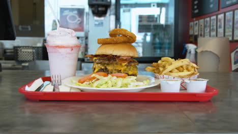 Woman-employee-at-a-burger-joint-pushes-a-full-tray-with-the-largest-burger-and-fries-and-shake-across-the-counter-for-the-customer-to-pick-up