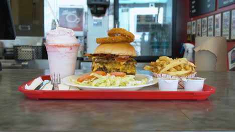 una empleada en una hamburguesería empuja una bandeja llena con la hamburguesa y papas fritas más grande y la sacude en el mostrador para que el cliente la recoja