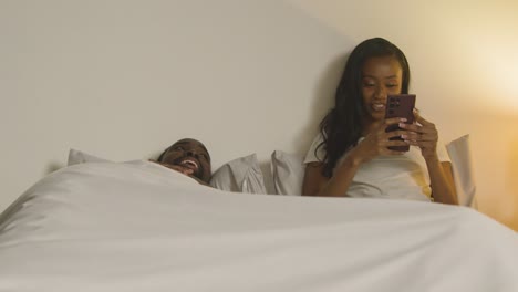 Pareja-En-Casa-Por-La-Noche-Acostada-En-La-Cama-Con-Una-Mujer-Mirando-El-Teléfono-Móvil-3