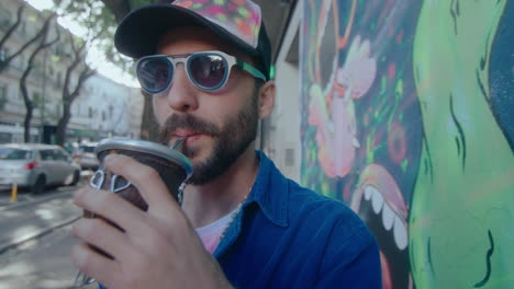 trendy street artist drinking from mug and posing by graffiti on the wall