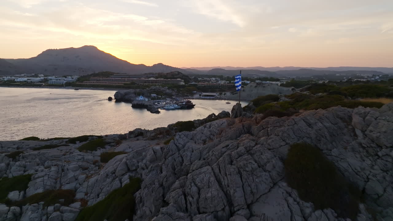 Drone flying past a Greece flag on a cliff, in front of Kolympia, sunset in Rodos