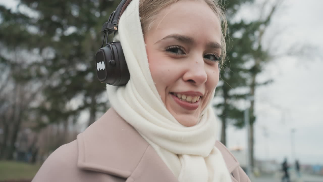 Donna al punto panoramico del parco con cuffie che usa un visore a moneta, avvolta in sciarpa bianca e cappotto beige, con un sorriso giocoso e un gesto della mano. Cielo coperto, viale alberato, ritratto ravvicinato cinematografico.