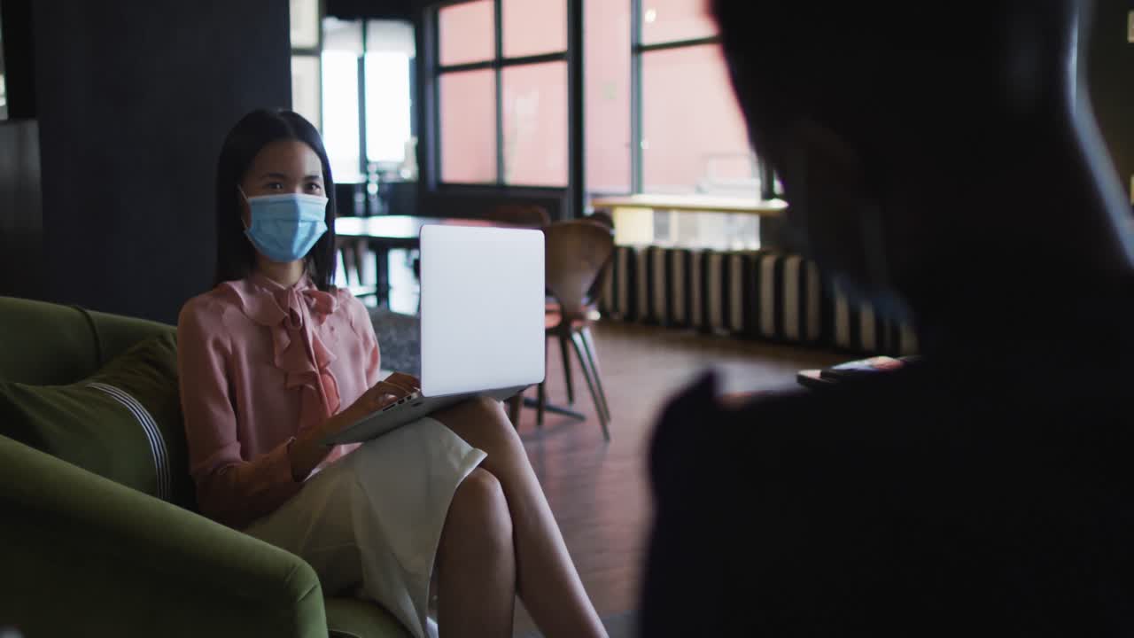 현대 사무실에서 소파 소파에 앉아서 노트북을 사용하는 얼굴 마스크를 착용 한 아시아 여성