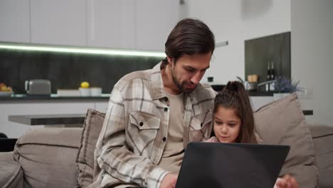 Ein-Brünetter-Mann-Mit-Stoppeln-Und-Beigefarbenem-Hemd-Kommuniziert-Mit-Seiner-Kleinen-Tochter,-Während-Er-Arbeitet-Und-Zeit-An-Einem-Grauen-Laptop-Auf-Dem-Sofa-In-Einem-Modernen-Apartment-Verbringt